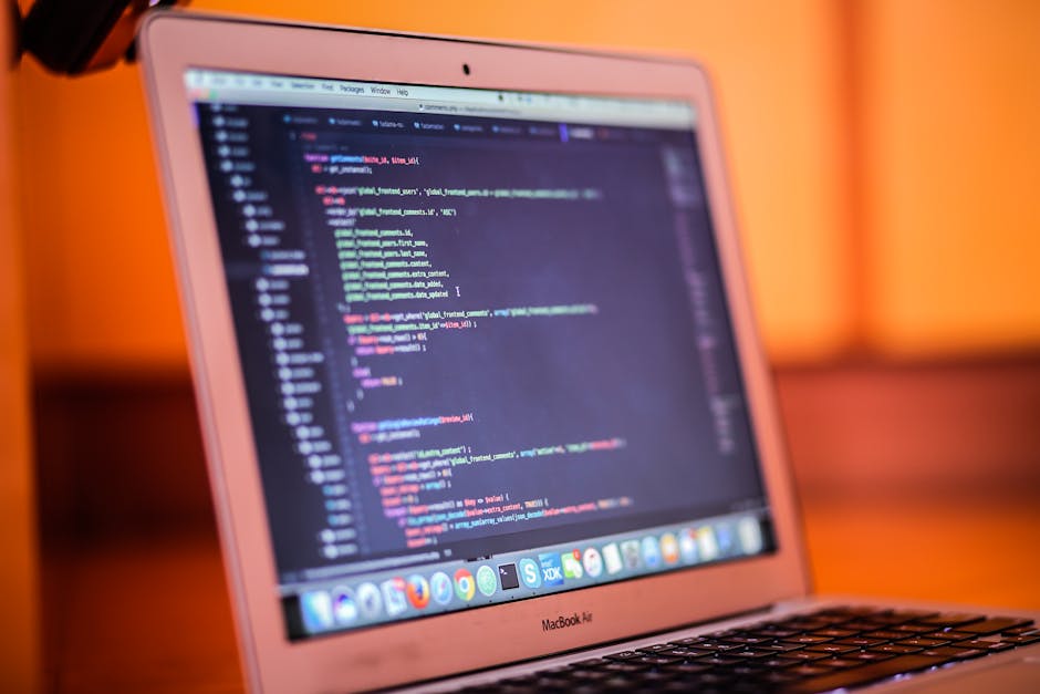 A close-up view of a MacBook Air laptop with a silver body, displaying a code editor filled with colorful programming syntax on the screen. The background is softly blurred with warm orange and brown 