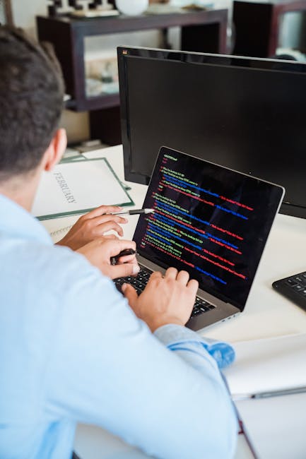 The image shows a person working at a desk with a laptop displaying lines of colorful computer code. The individual, seen from behind, is focused on the screen and holding a pen. On the desk, there is